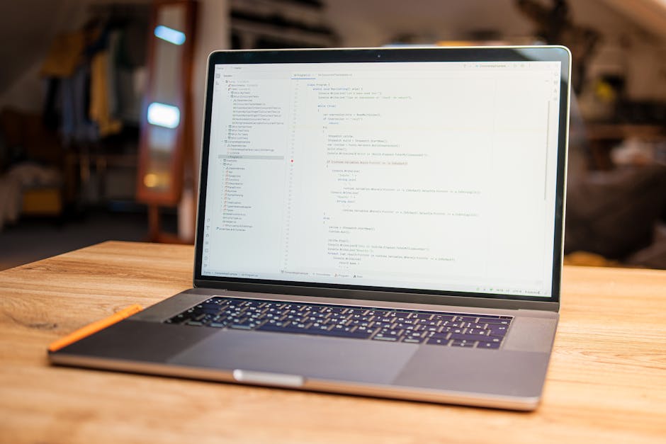 A person’s hands are visible typing on a laptop keyboard with a dark screen displaying lines of colorful code, placed on a black table. The background is slightly blurred, showing an outdoor setting w