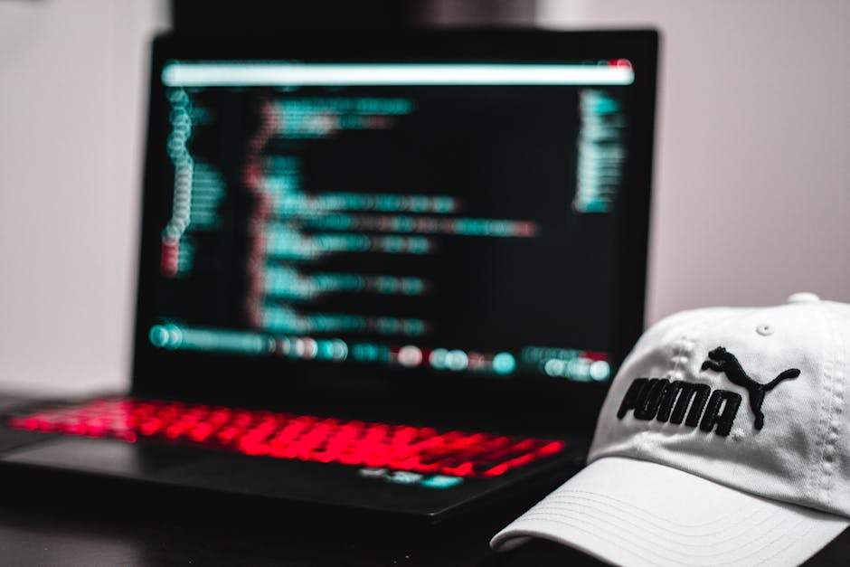 The image features a black laptop with a red backlit keyboard placed on a surface, displaying blurred lines of code on the screen with a neon blue and pink glow. Next to the laptop, there is a white b